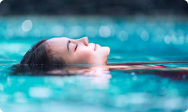 A woman floats on her back in a swimming pool with her eyes closed, appearing relaxed.