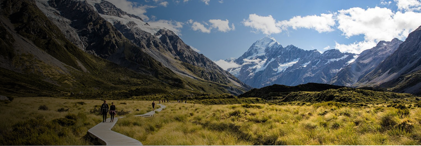 People walking on a wooden boardwalk through grassy fields, with tall snow-capped mountains and a partly cloudy sky in the background.