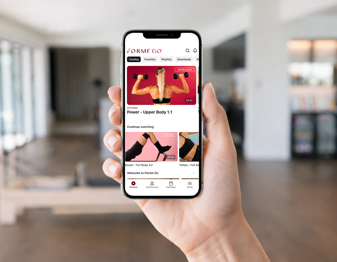 A person holds a smartphone displaying a fitness app with workout videos in a modern, bright living room.
